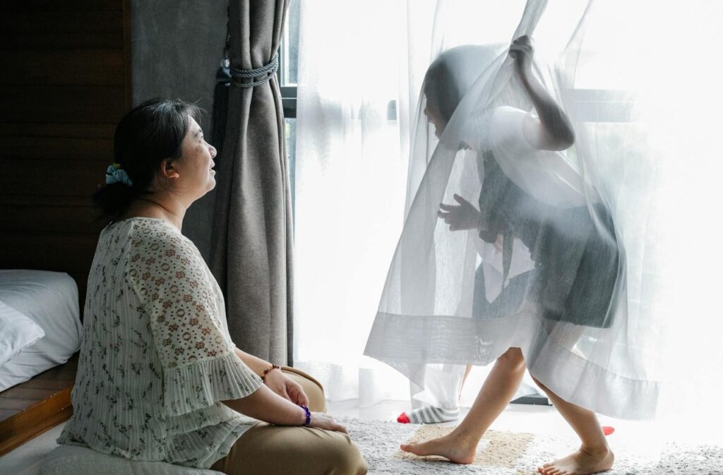 一位女子坐在窗邊微笑仰望，赤腳的孩子躲在白色紗簾後玩耍，午後自然光灑入臥室的溫馨親子時刻。