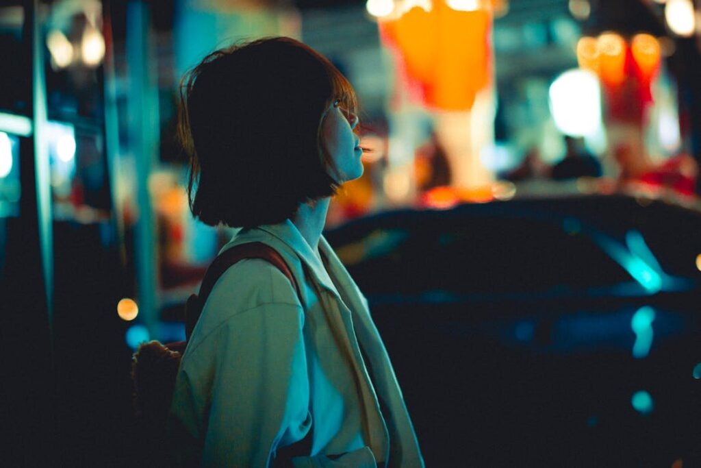 一名留著短髮的女子側臉望向前方，站在夜晚的街頭，背景是模糊的橘色與藍綠色霓虹光斑與一輛車的輪廓，整體呈現電影感的城市夜色。